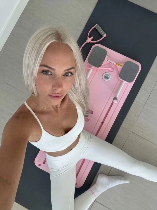 A young woman in white gym wear posing with an EasyLean pink foldable pilates reformer, ideal for home body sculpting and postpartum recovery.