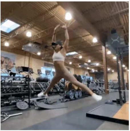 A fit woman in a white sports bra exploding into a burpee with arm-spreading jump mid-air in the gym to break a home fitness plateau