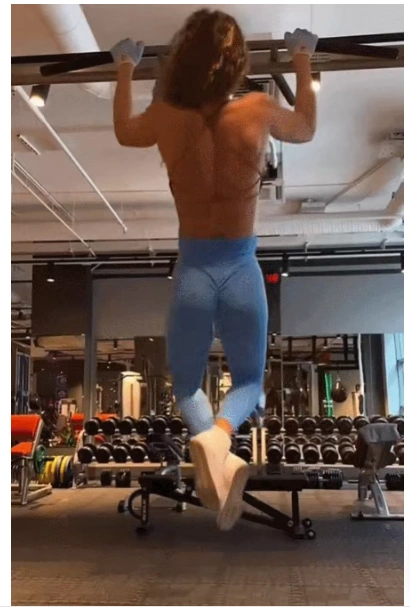 A fit woman with a defined back performing a strict pull-up in the gym to test her relative strength and break a home fitness plateau