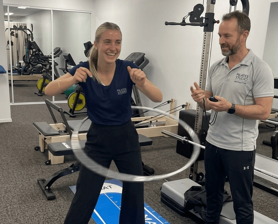 smart hula hoop vs. Traditional Hula Hoops: The Ultimate 2026 Comparison (No Bruises, No Drops!) 2 A smiling young woman in a fitness studio performs a traditional weighted hula hoop rotation challenge, while a personal trainer standing next to her tracks the workout time with a stopwatch.